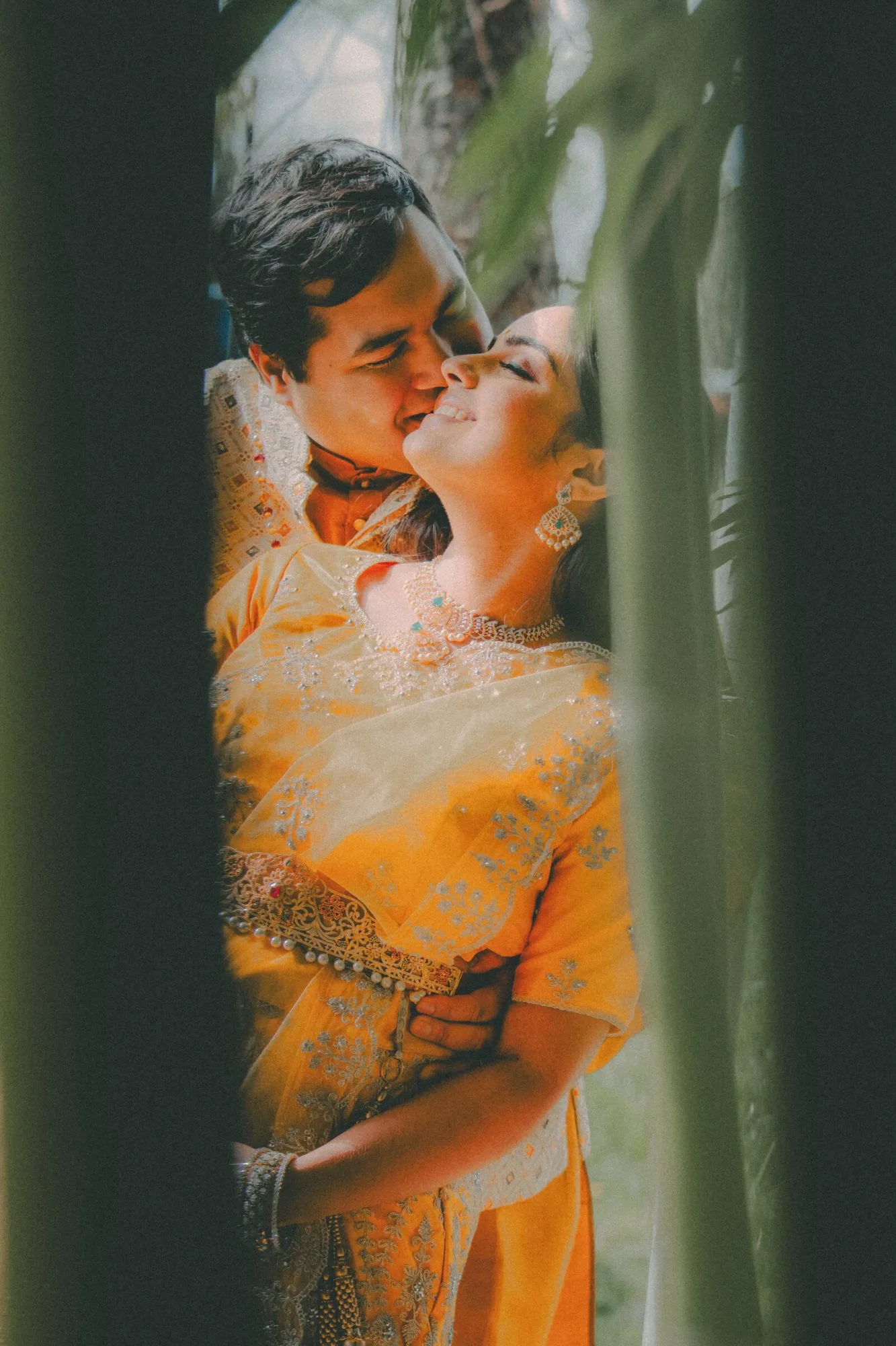 A couple in marigold and gold, photographed close between palm leaves at their celebration