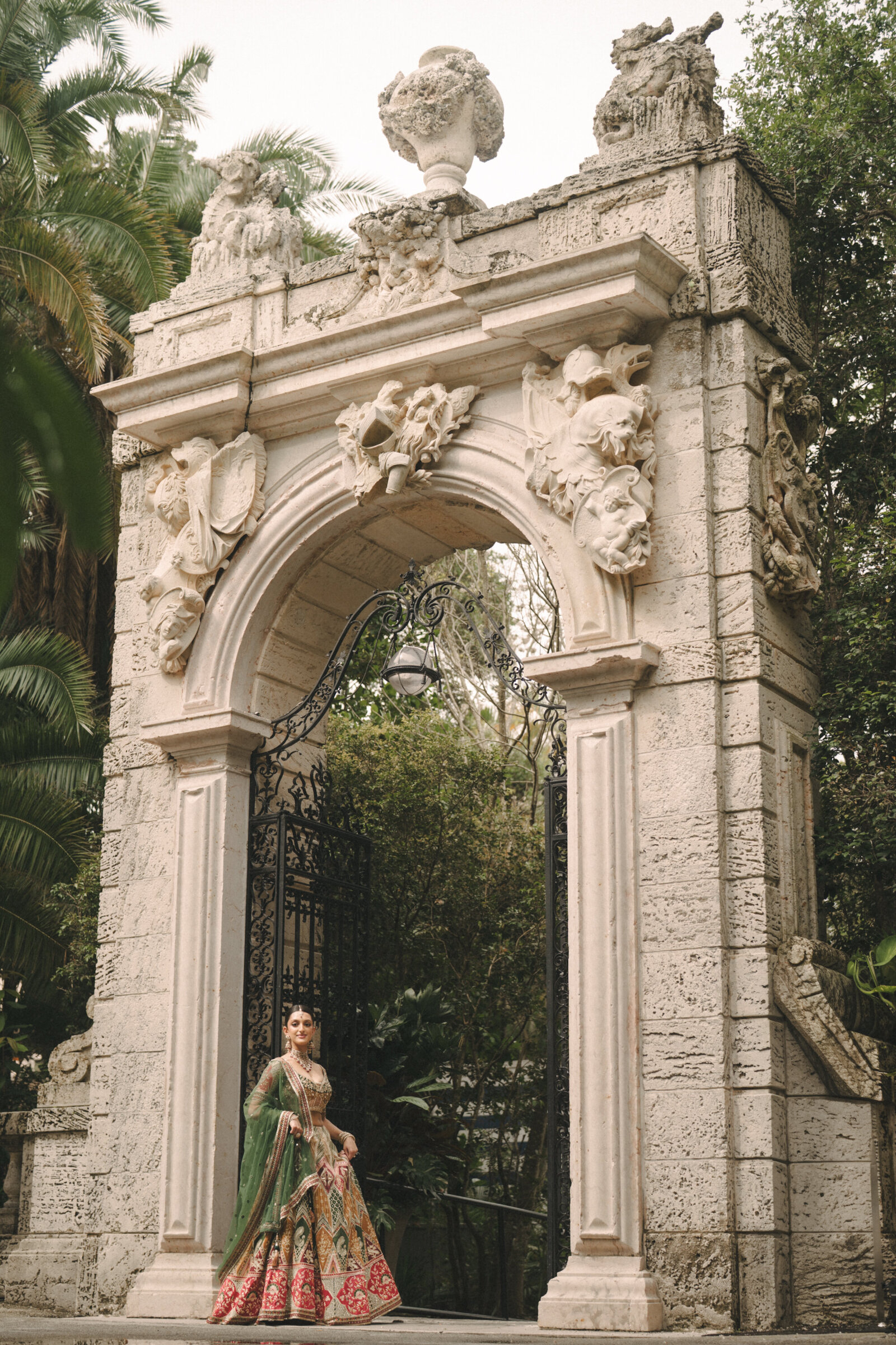 A bride in a green and red lehenga beneath a carved stone arch