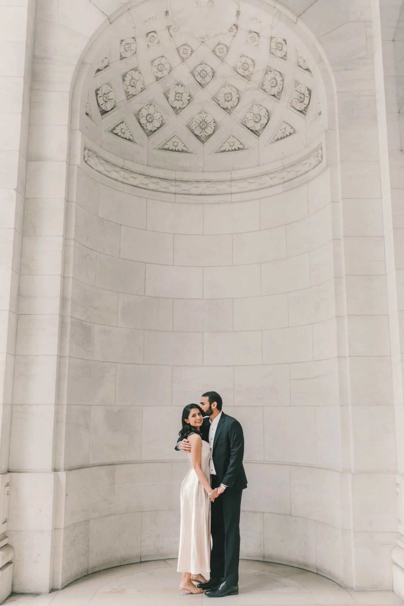 A couple beneath a carved white stone arch, the groom kissing the bride's temple