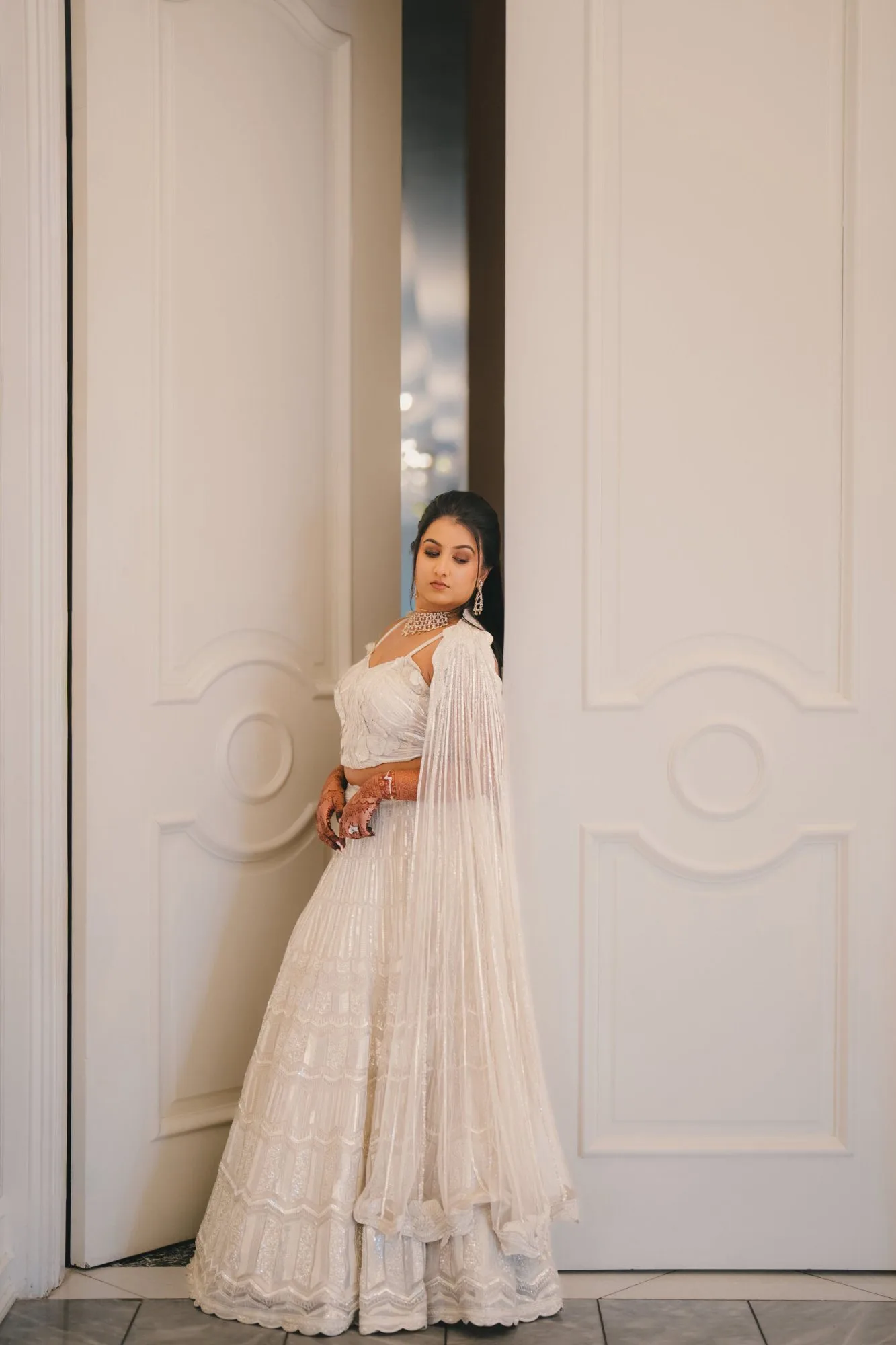 A bride in an ivory embellished lehenga with deep henna on her hands, leaning against tall white doors