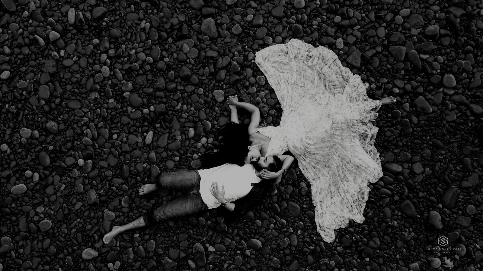 An aerial black and white frame of a couple lying on a pebble shore