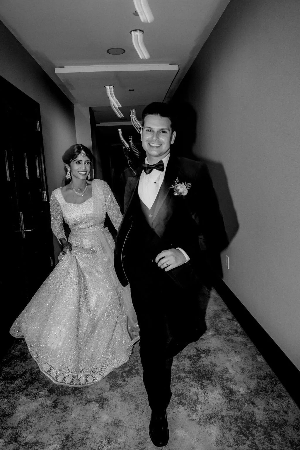 A bride in an embellished lehenga and a groom in a tuxedo hurrying down a corridor, laughing