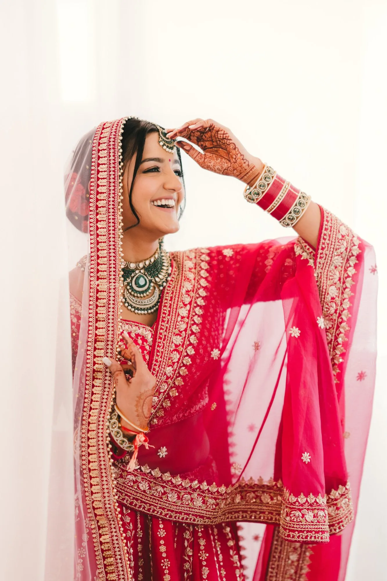 A bride in a red lehenga and chooda laughing as she lifts her dupatta in soft window light