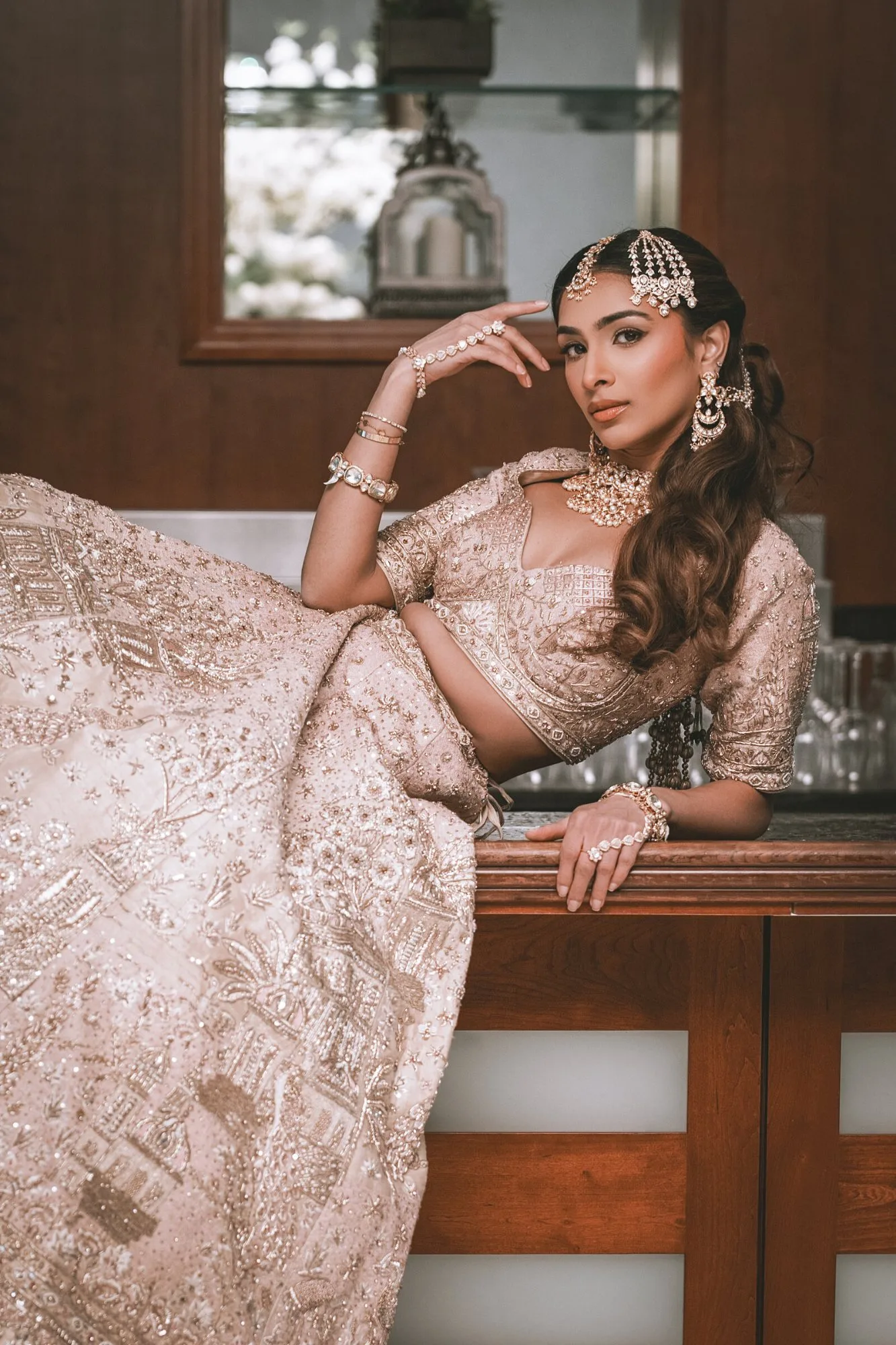 A bride in a blush embroidered lehenga, resting against a wooden bar with her jewellery catching the light