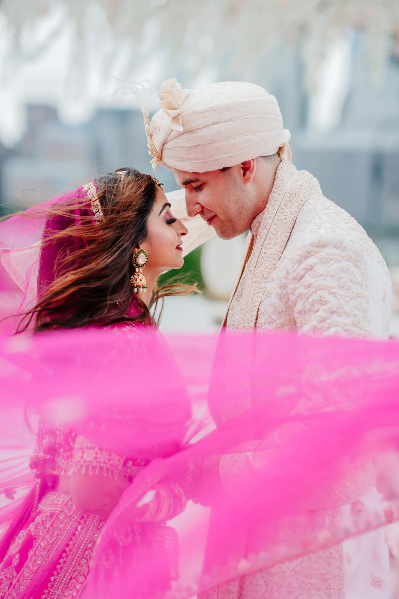 A bride and groom beneath a bright pink dupatta, the city soft behind them